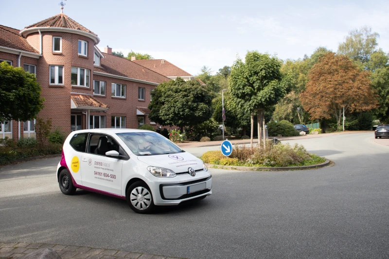 Pflegefahrzeug vor Seniorenheim in Wohngebiet, umgeben von Bäumen und moderner Architektur.