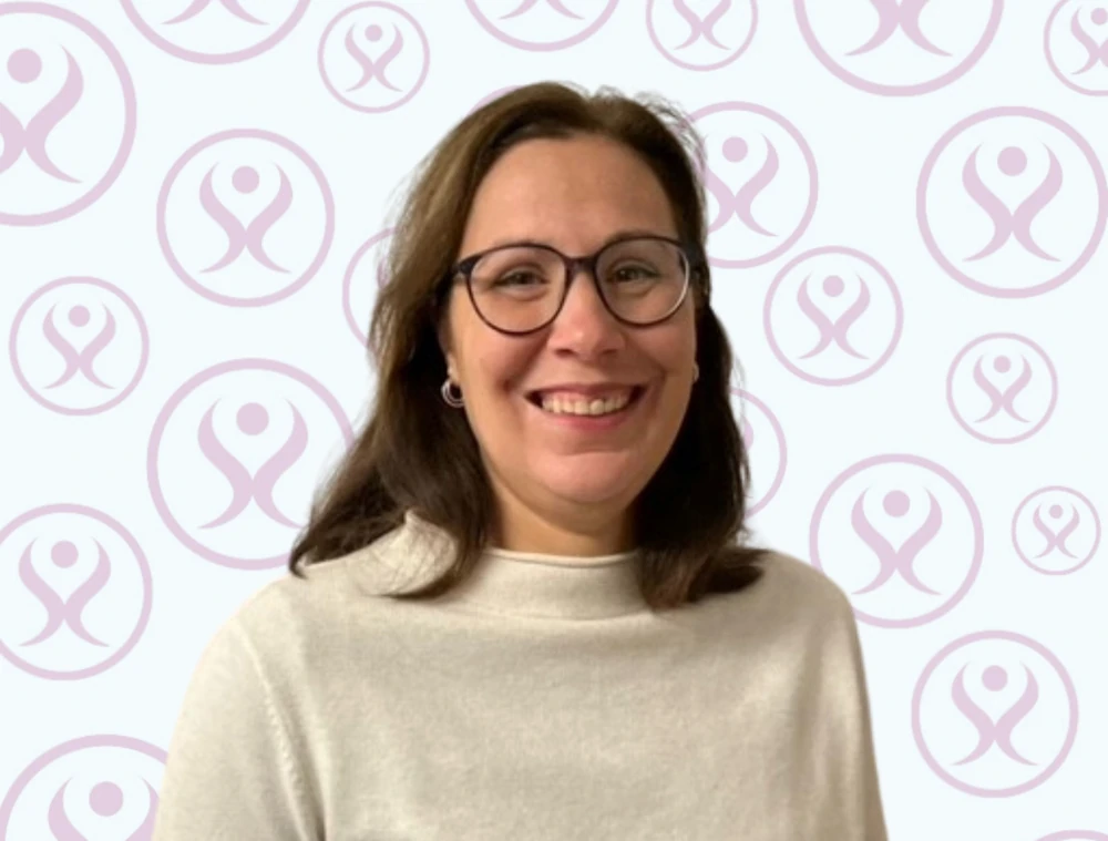 Frau mit Brille und weißem Pullover lächelt vor hellem Hintergrund mit Firmenlogo.